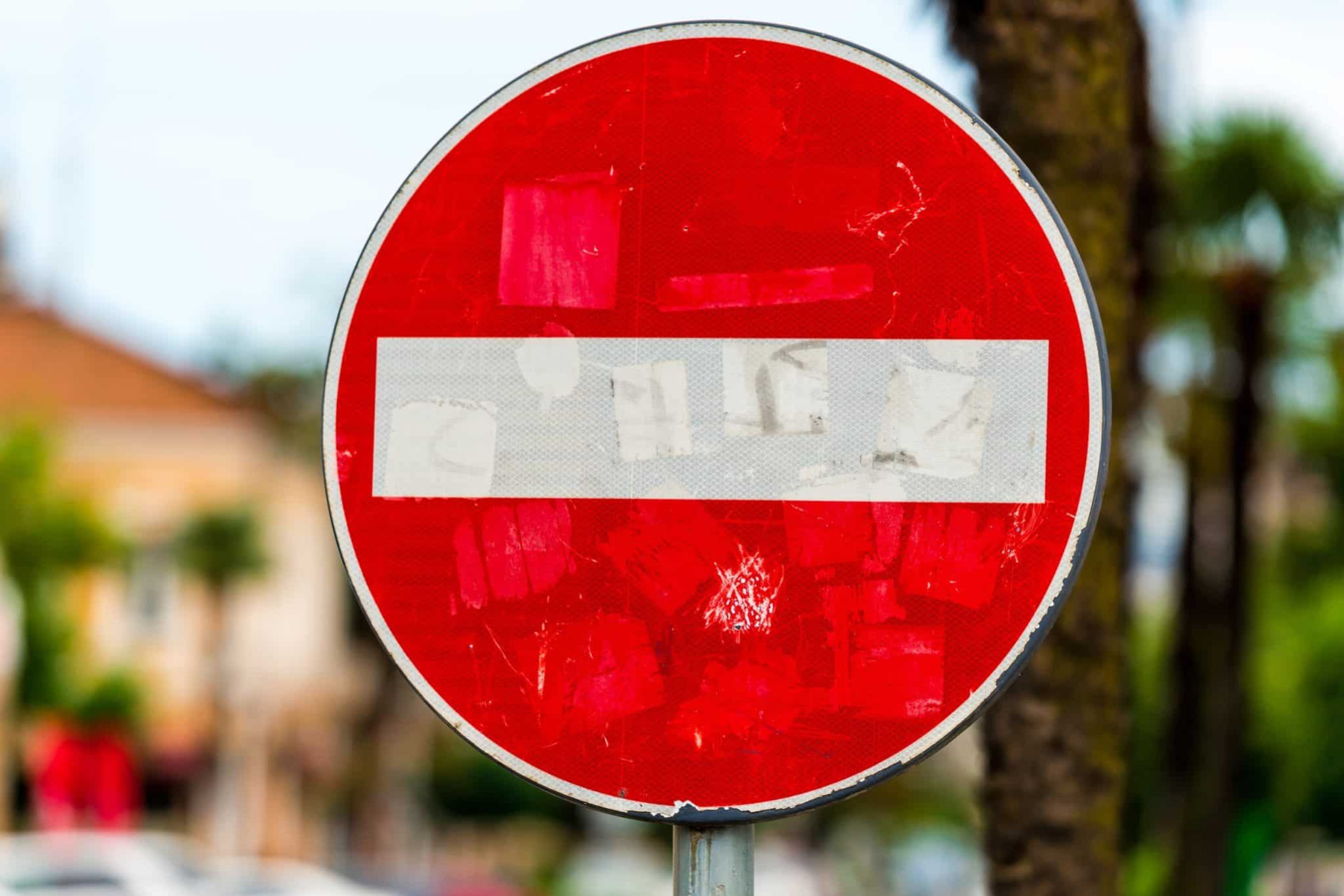 A red and white "Do Not Enter" traffic sign, symbolizing common workers’ comp mistakes, is faded and covered with worn stickers, positioned outdoors with trees and buildings in the blurred background.