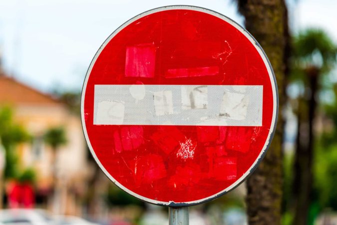 A red and white "Do Not Enter" traffic sign, symbolizing common workers’ comp mistakes, is faded and covered with worn stickers, positioned outdoors with trees and buildings in the blurred background.