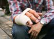 A person wearing a plaid shirt sits outdoors with their left hand wrapped in a bandage—possibly considering workers’ compensation or curious about case settlement time after a recent injury.