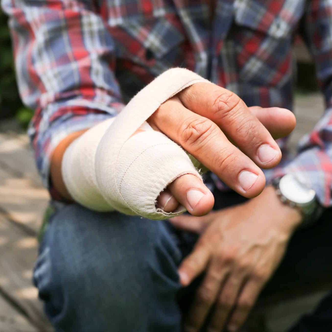 A person wearing a plaid shirt sits outdoors with their left hand wrapped in a bandage—possibly considering workers’ compensation or curious about case settlement time after a recent injury.