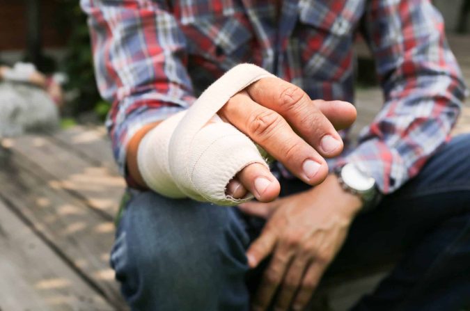 A person wearing a plaid shirt sits outdoors with their left hand wrapped in a bandage—possibly considering workers’ compensation or curious about case settlement time after a recent injury.