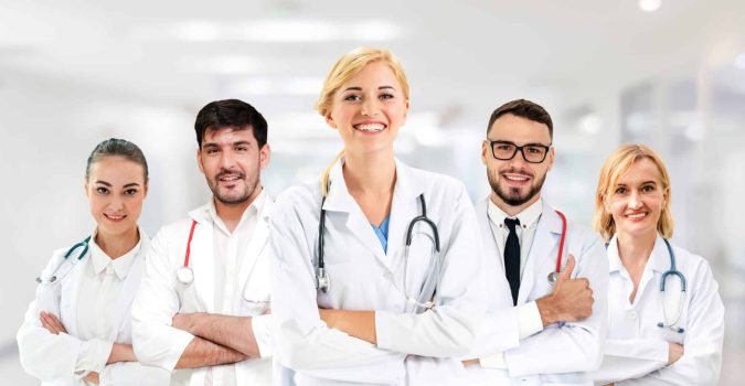 Five medical professionals in white lab coats stand together in a hospital hallway, smiling and looking at the camera—ready to treat patients as trusted Lansing Workers’ Comp Doctors.
