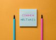 A blue sticky note with the words "COMMON MISTAKES" written on it, placed between a blue pen and a pink pen on an orange background—a helpful reminder for those in Michigan applying for Social Security Disability.