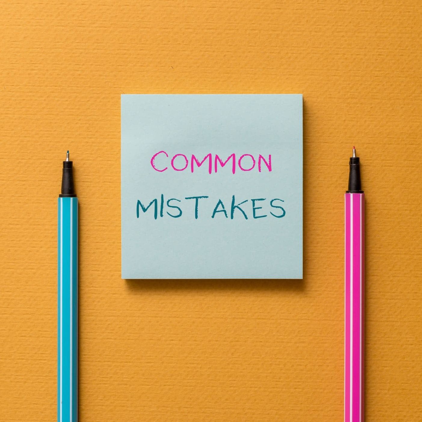 A blue sticky note with the words "COMMON MISTAKES" written on it, placed between a blue pen and a pink pen on an orange background—a helpful reminder for those in Michigan applying for Social Security Disability.