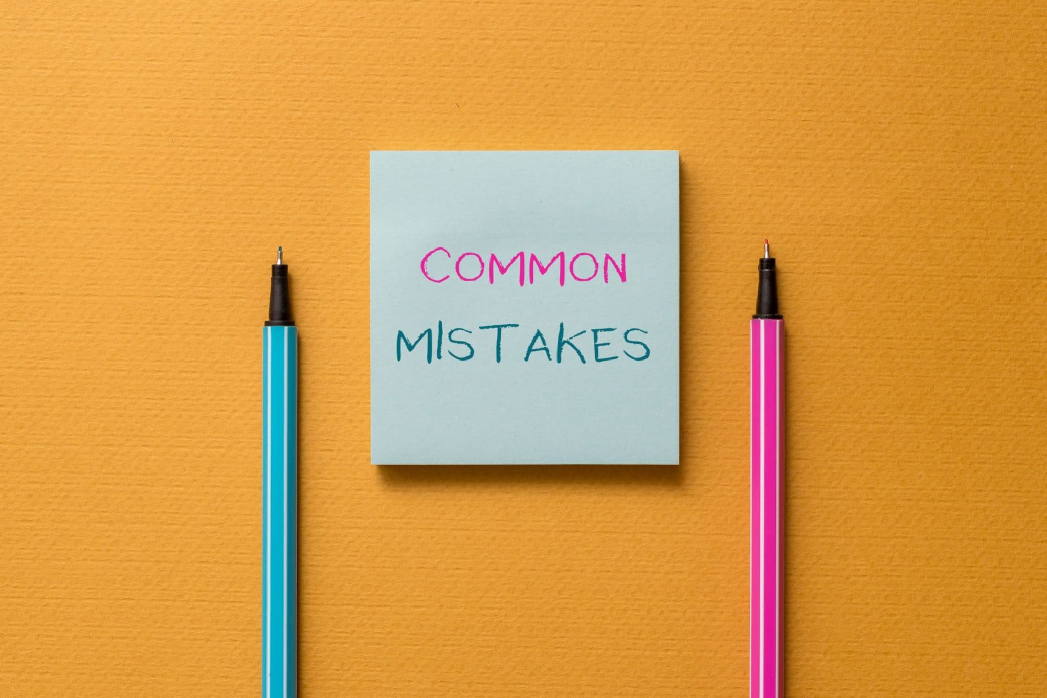 A blue sticky note with the words "COMMON MISTAKES" written on it, placed between a blue pen and a pink pen on an orange background—a helpful reminder for those in Michigan applying for Social Security Disability.
