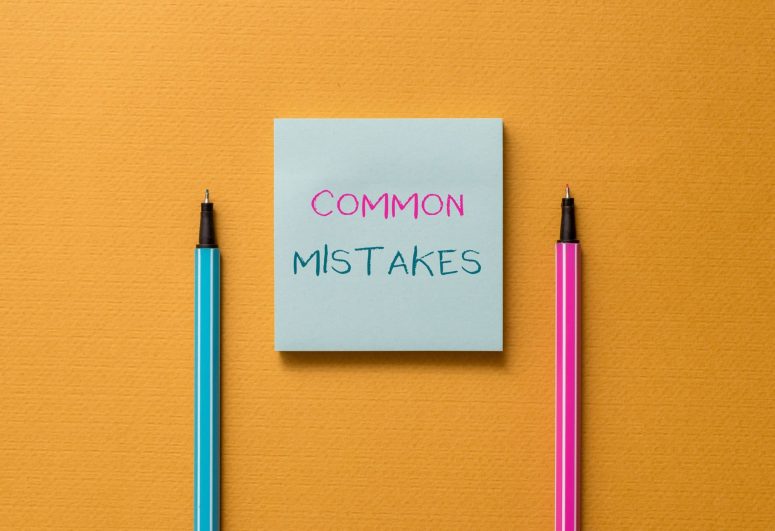 A blue sticky note with the words "COMMON MISTAKES" written on it, placed between a blue pen and a pink pen on an orange background—a helpful reminder for those in Michigan applying for Social Security Disability.