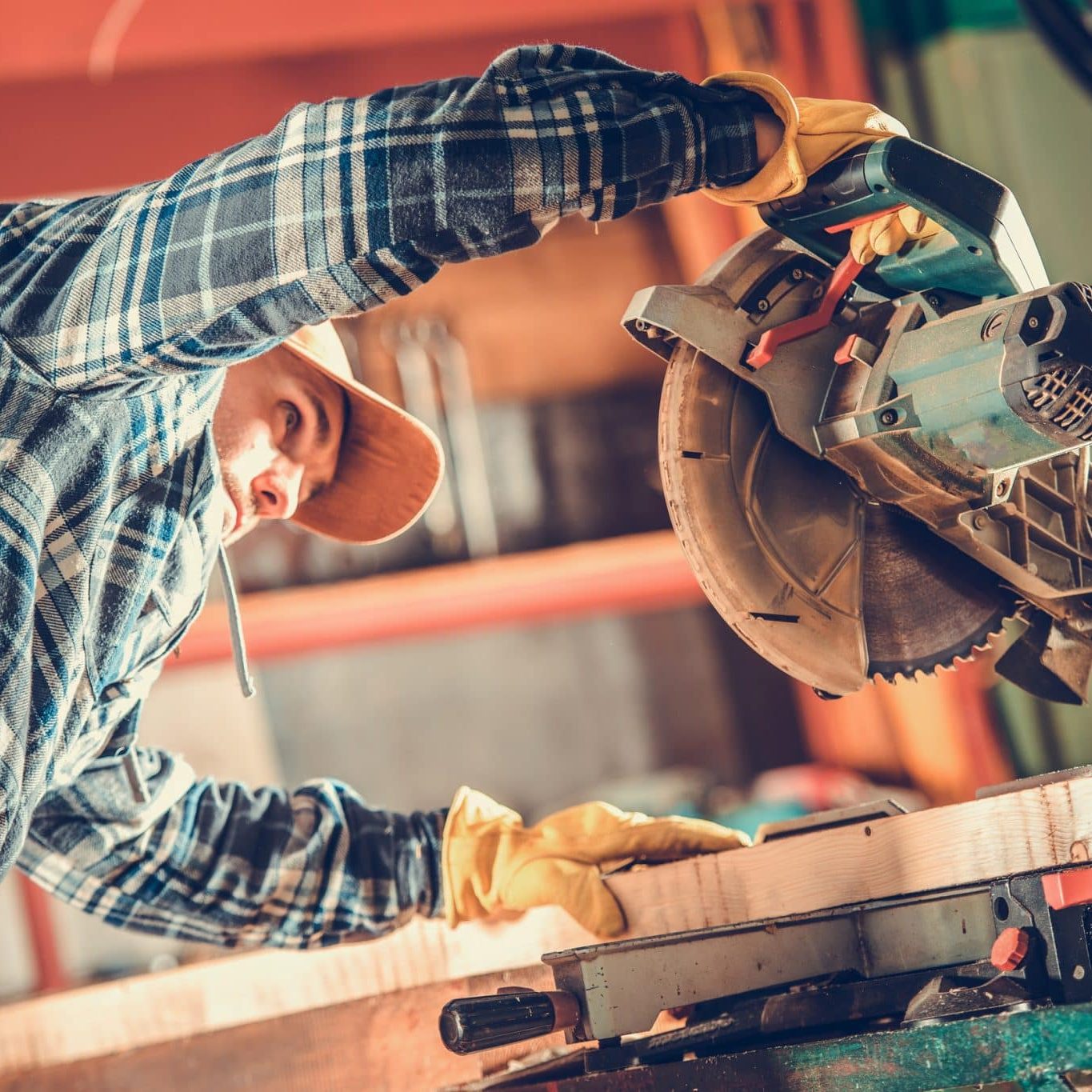 A person wearing gloves and a plaid shirt uses a circular saw to cut a wooden board in a workshop.