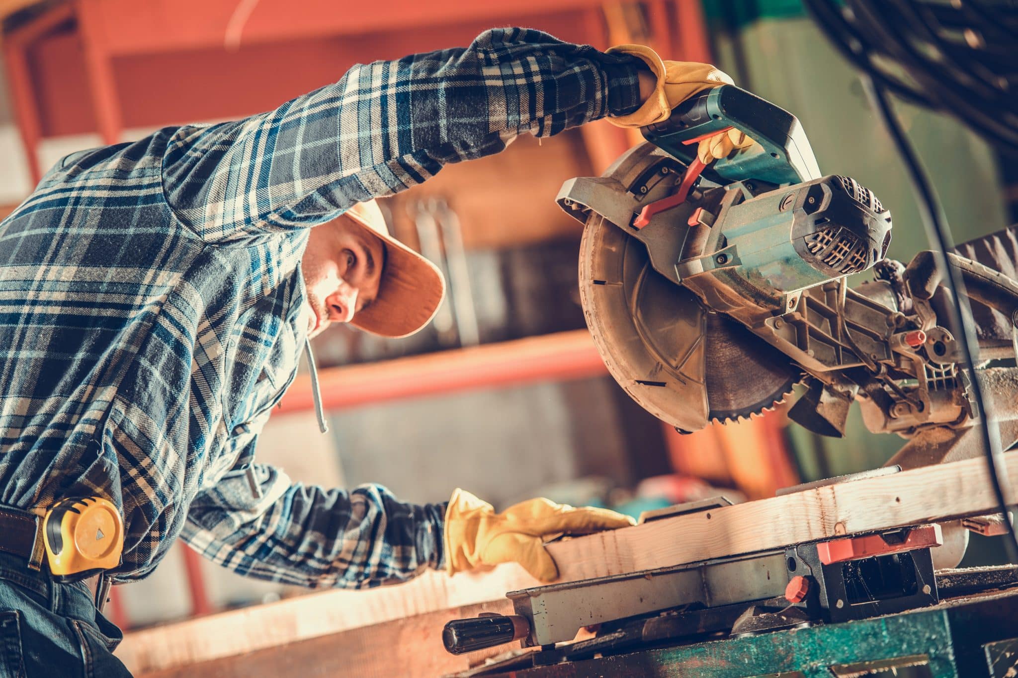 A person wearing gloves and a plaid shirt uses a circular saw to cut a wooden board in a workshop.