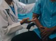 A doctor in a white coat discusses Workers’ Comp with a seated patient in a medical gown, the doctor's hand gesturing as they talk—offering insights similar to an expert Survival Guide.