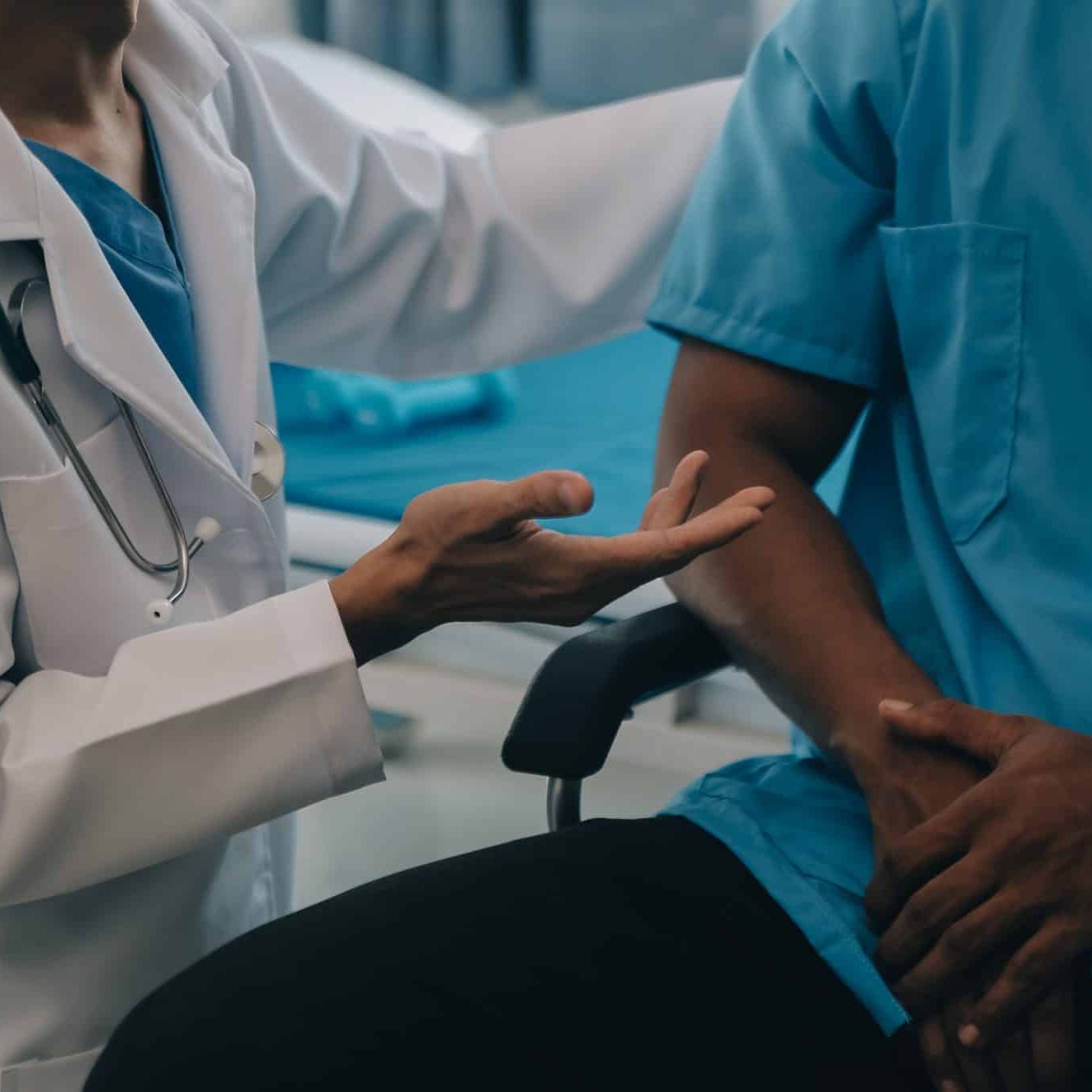 A doctor in a white coat discusses Workers’ Comp with a seated patient in a medical gown, the doctor's hand gesturing as they talk—offering insights similar to an expert Survival Guide.