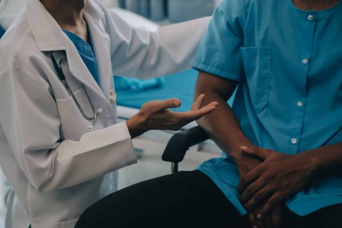 A doctor in a white coat discusses Workers’ Comp with a seated patient in a medical gown, the doctor's hand gesturing as they talk—offering insights similar to an expert Survival Guide.