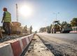 Road construction site with workers in safety vests and hard hats, heavy machinery, and bright sunlight in the background.