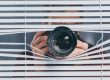A person with a camera peers through partially open window blinds, aiming the lens outward.