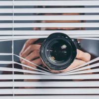A person with a camera peers through partially open window blinds, aiming the lens outward.