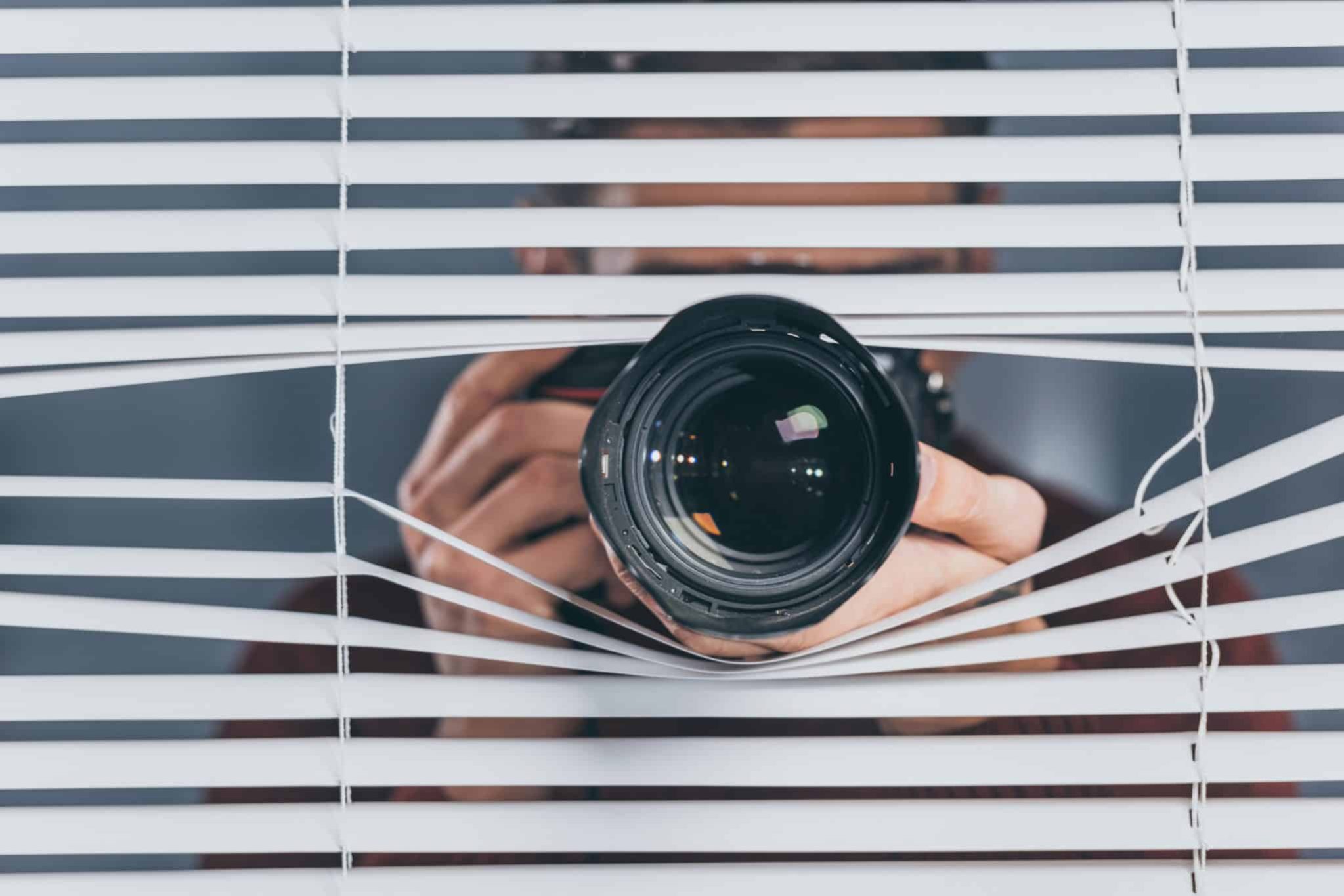 A person with a camera peers through partially open window blinds, aiming the lens outward.