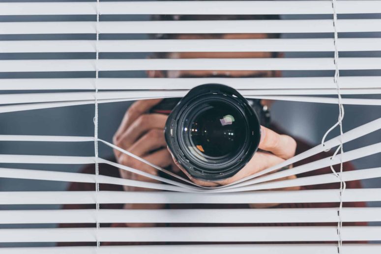 The Hidden Tactics Insurance Companies Use in Wrongful Death Claims A person with a camera peers through partially open window blinds, aiming the lens outward.