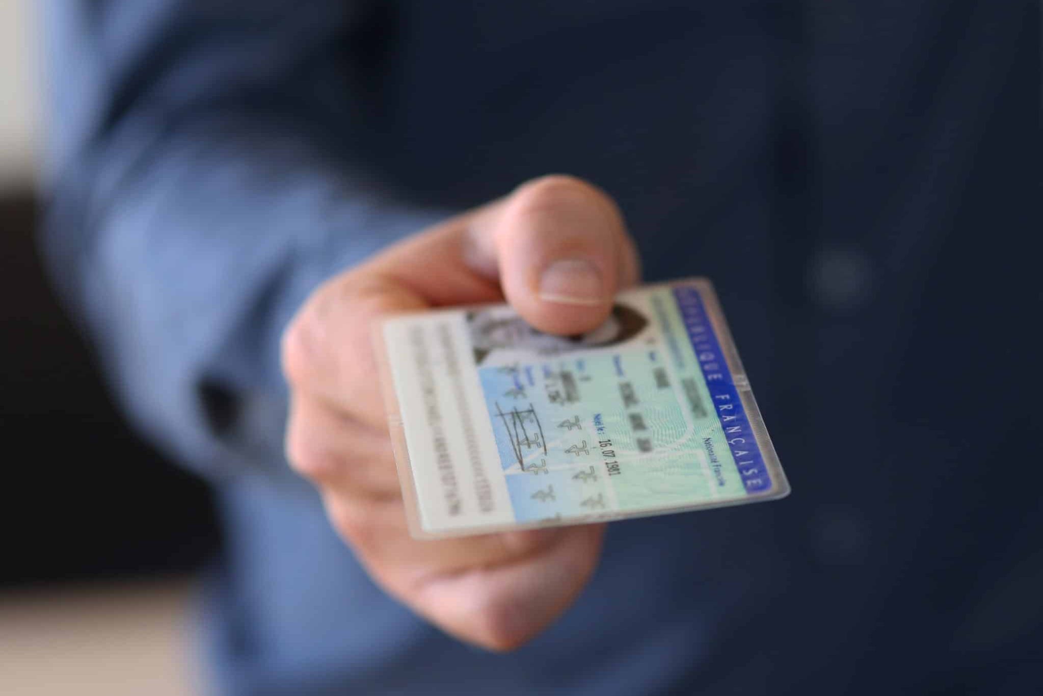 A person in a blue shirt holds out a French identification card, showing the front side with text and a photo visible.