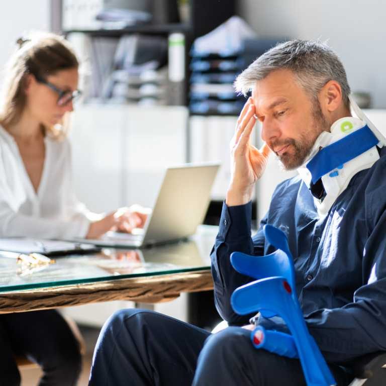 A man with a neck brace and crutches sits at a desk looking concerned, while a woman works on a laptop in the background.