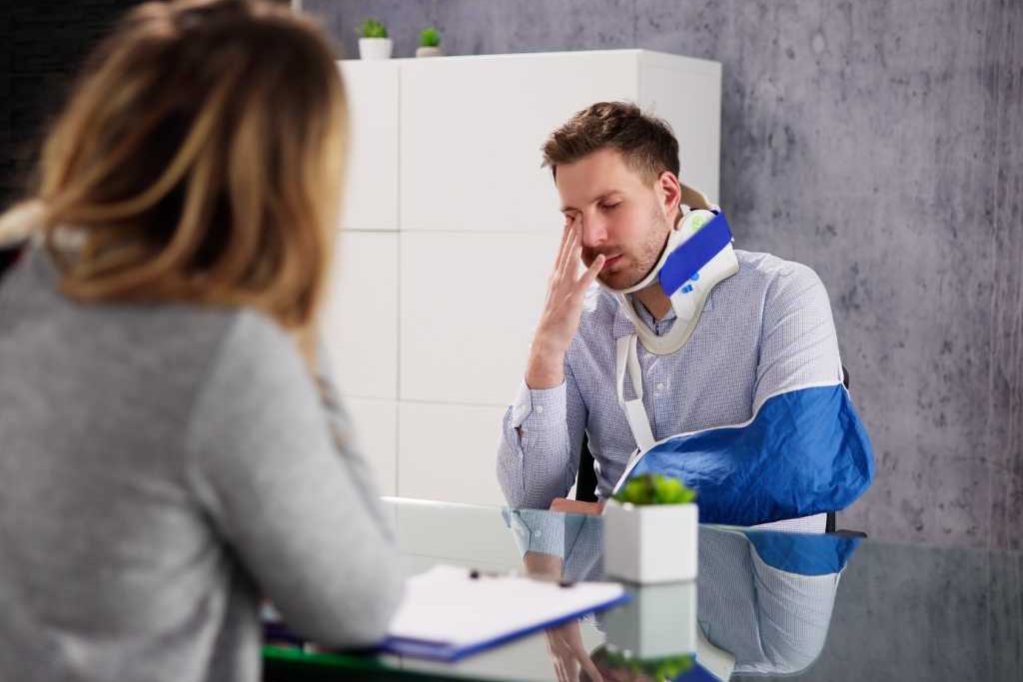A man with a neck brace and arm sling sits at a desk across from a woman, holding his head with his hand in a modern office setting.