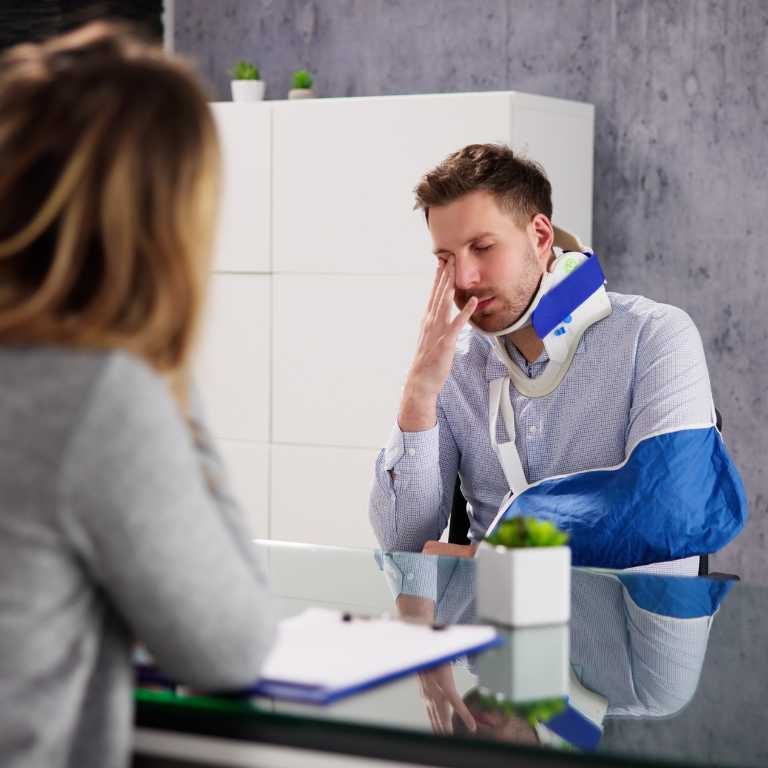 A man with a neck brace and arm sling sits at a desk across from a woman, holding his head with his hand in a modern office setting.