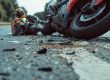 A motorcycle lies on its side amid debris on a road, with a person in a helmet and protective gear sitting nearby.