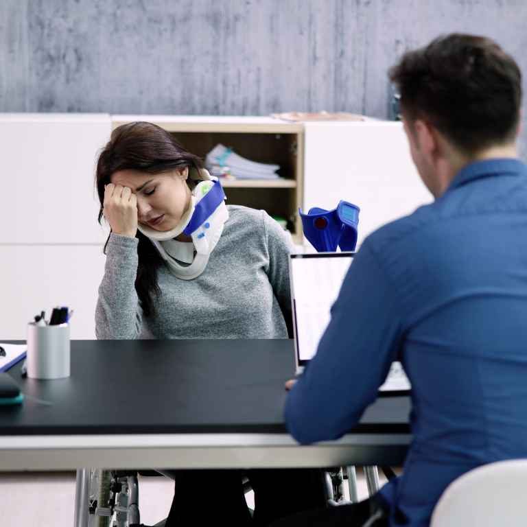 A woman wearing a neck brace sits at a desk, holding her head in discomfort, while a man in a blue shirt sits across from her, possibly in a meeting or consultation.