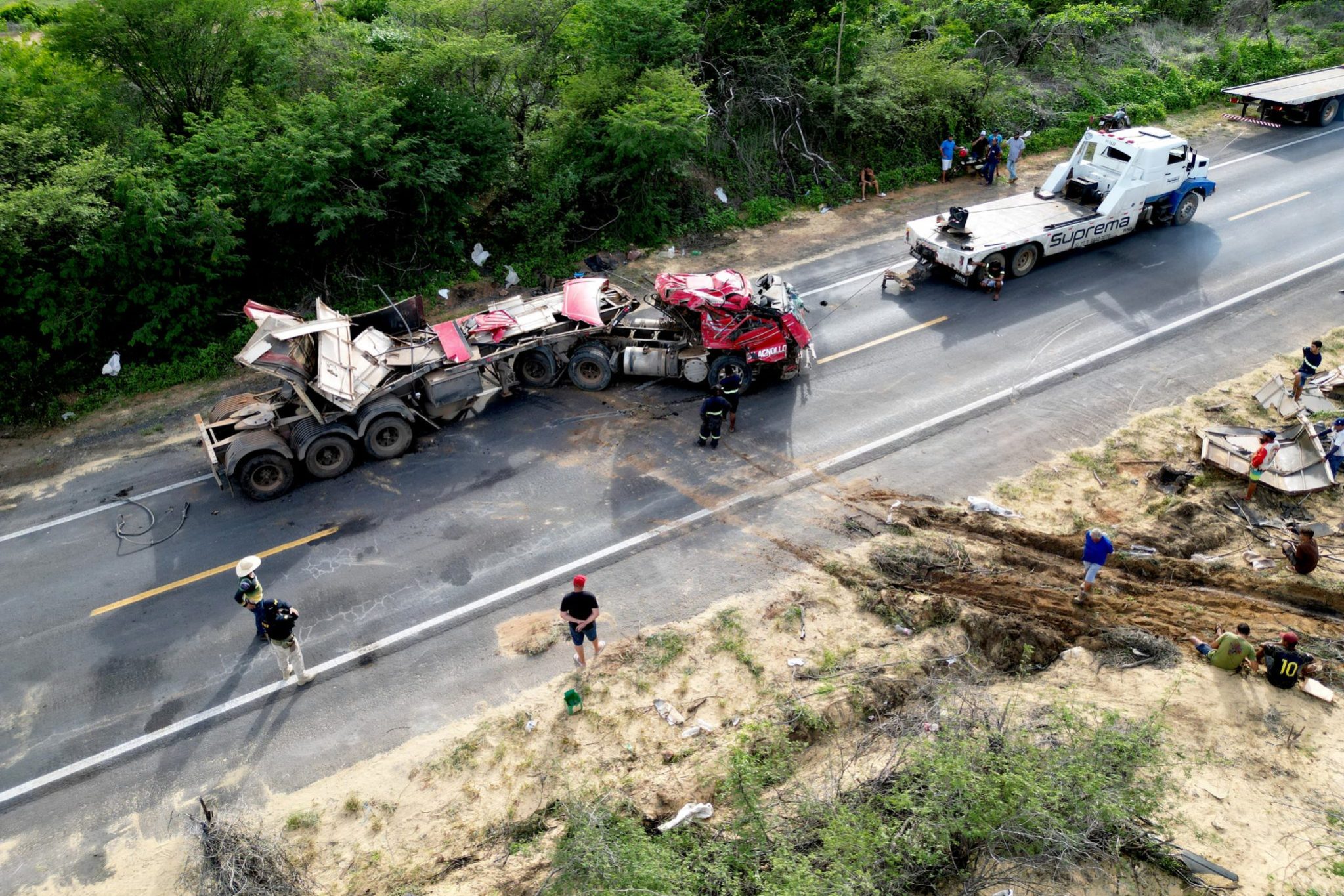 Aerial view of a damaged semi-truck and tow truck on a two-lane road, with workers and debris scattered along the roadside.