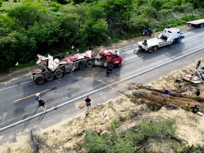 Aerial view of a damaged semi-truck and tow truck on a two-lane road, with workers and debris scattered along the roadside.