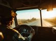 A person wearing a cap drives a vehicle down an empty rural road at sunset, as seen from inside the vehicle.