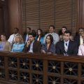Twelve people sit in a jury box inside a courtroom, appearing attentive and focused, with wood paneling and a window behind them.