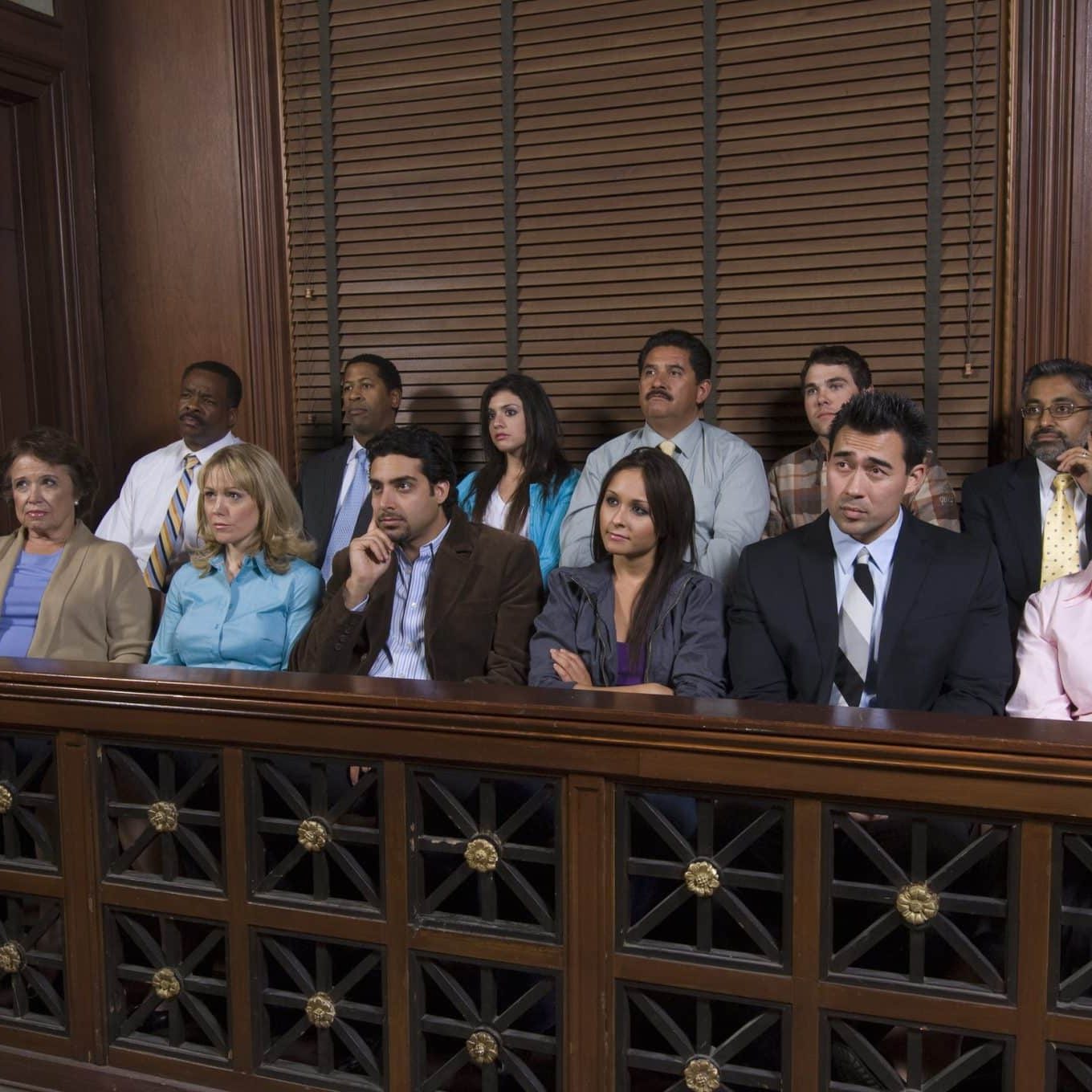 Lansing Juries Twelve people sit in a jury box inside a courtroom, appearing attentive and focused, with wood paneling and a window behind them.