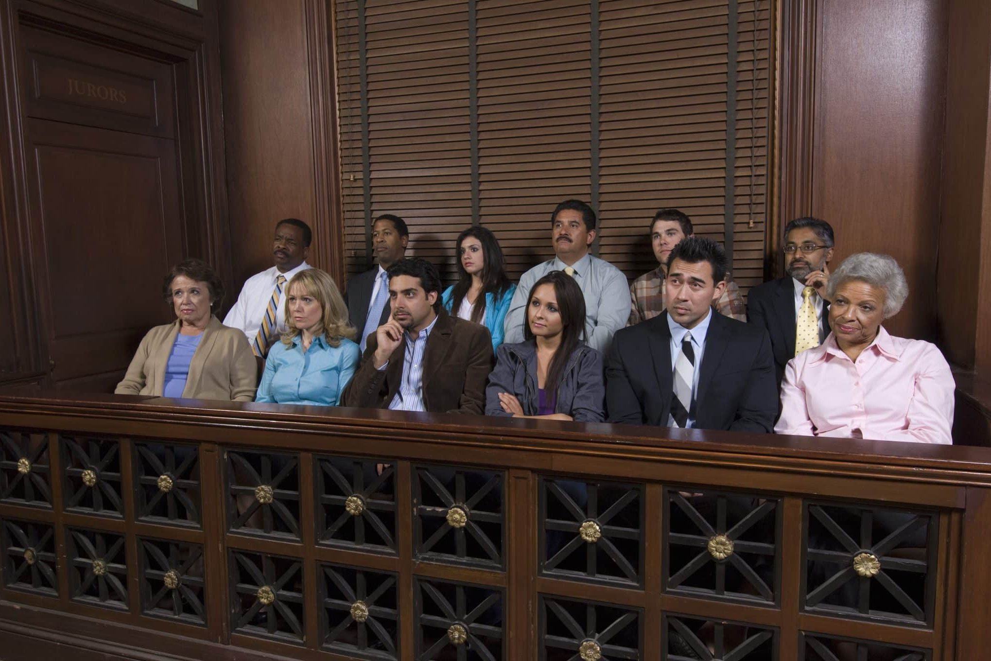 Lansing Juries Twelve people sit in a jury box inside a courtroom, appearing attentive and focused, with wood paneling and a window behind them.