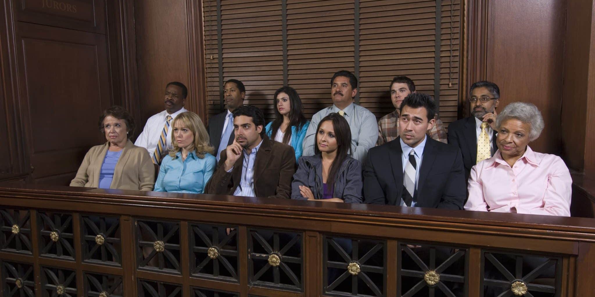 Lansing Juries Twelve people sit in a jury box inside a courtroom, appearing attentive and focused, with wood paneling and a window behind them.