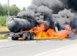 A tanker truck on a highway is engulfed in flames, producing thick black smoke that rises into the sky.
