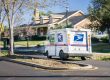 A USPS mail truck is parked on a suburban street in front of a house with a lawn and trees on a sunny day.