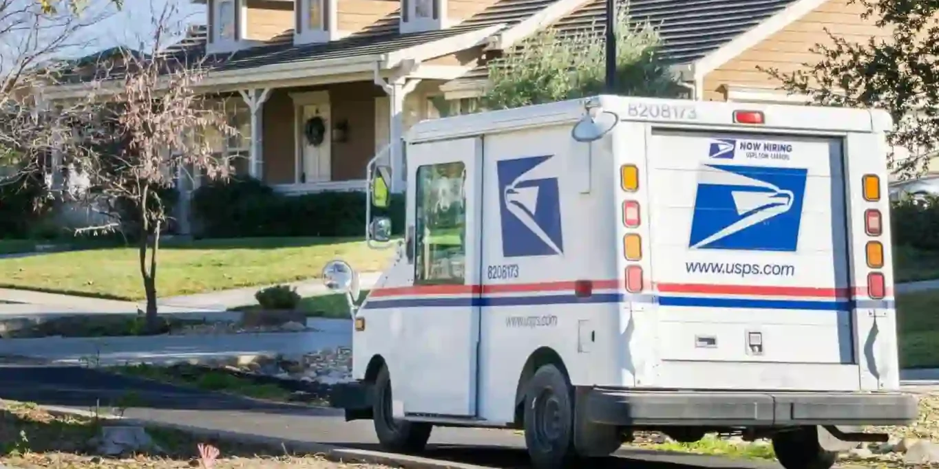 A USPS mail truck is parked on a residential street in front of a tan house with a porch and well-kept lawn. A USPS mail truck is parked on a residential street in front of a tan house with a porch and well-kept lawn.