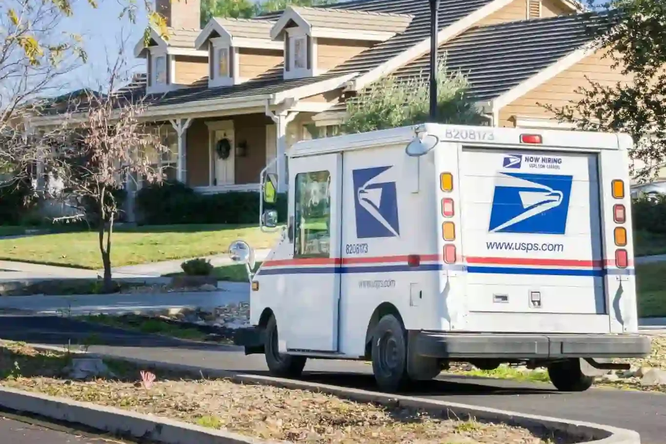 A USPS mail truck is parked on a residential street in front of a tan house with a porch and well-kept lawn.