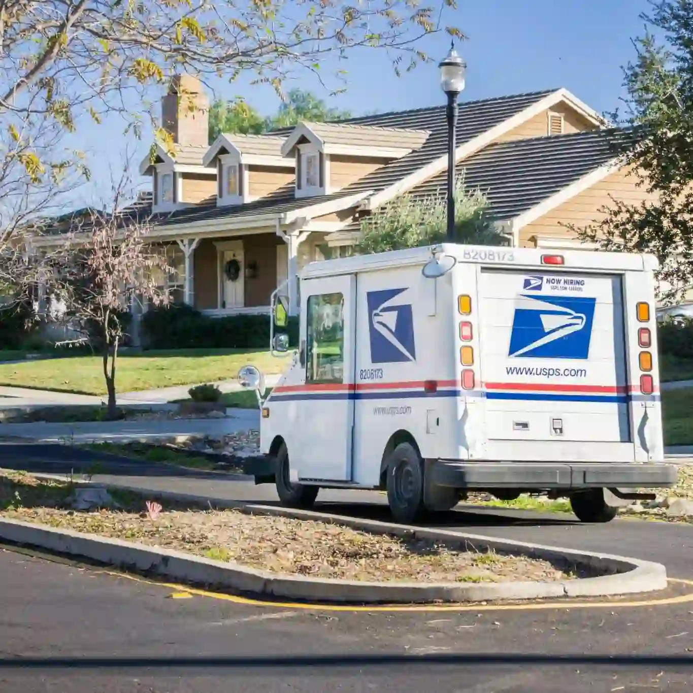 A USPS mail truck is parked on a residential street in front of a tan house with a porch and well-kept lawn.