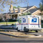 A USPS mail truck is parked on a suburban street in front of a house with a lawn and trees on a sunny day.