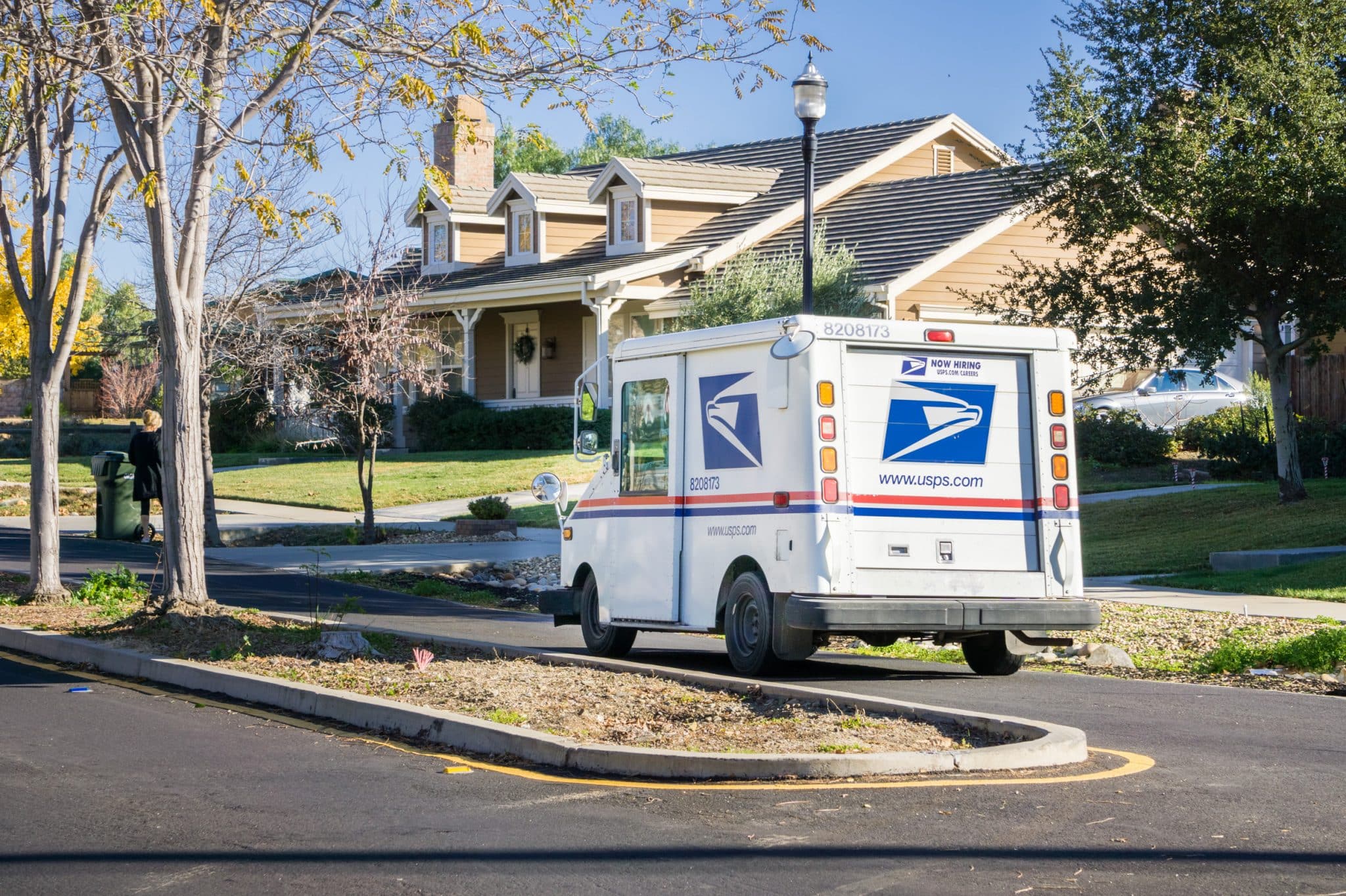 A USPS mail truck is parked on a suburban street in front of a house with a lawn and trees on a sunny day.