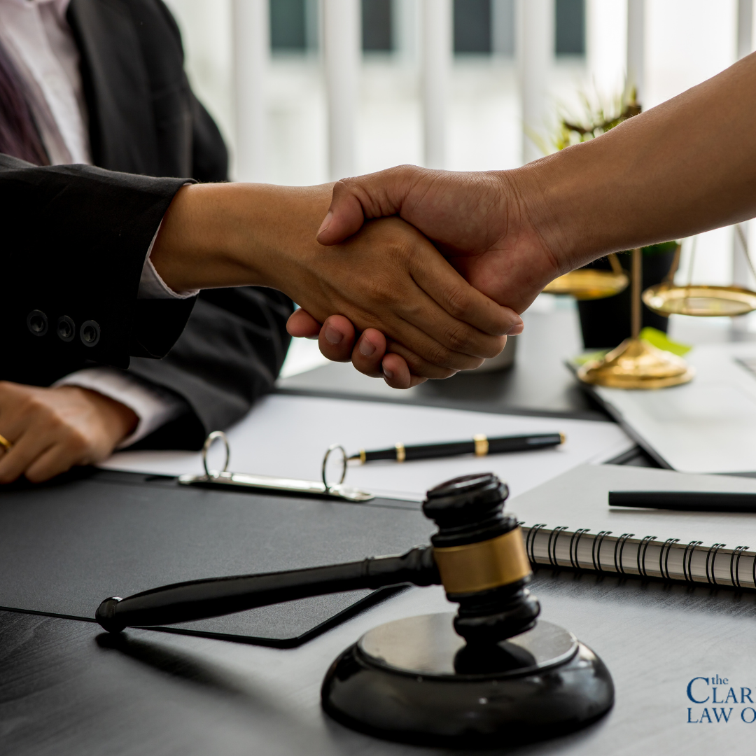 Two people shaking hands across a desk with legal documents, a gavel, and office supplies visible; Clark Law Office logo in the corner. Two people shaking hands across a desk with legal documents, a gavel, and office supplies visible; Clark Law Office logo in the corner.