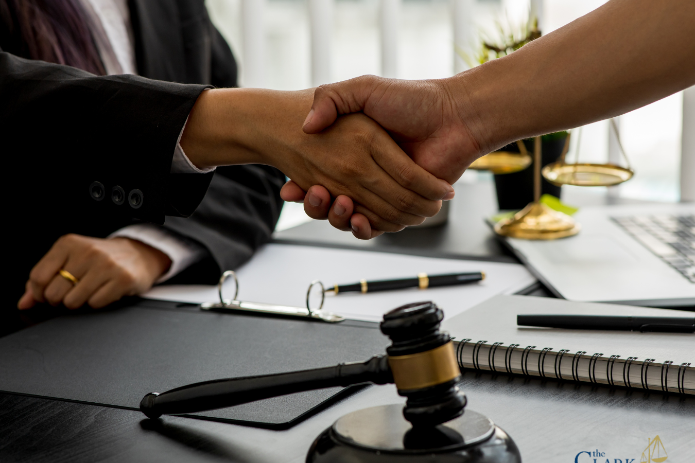 Two people shaking hands across a desk with legal documents, a gavel, and office supplies visible; Clark Law Office logo in the corner.
