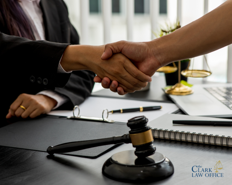 Two people shaking hands across a desk with legal documents, a gavel, and office supplies visible; Clark Law Office logo in the corner. Two people shaking hands across a desk with legal documents, a gavel, and office supplies visible; Clark Law Office logo in the corner.