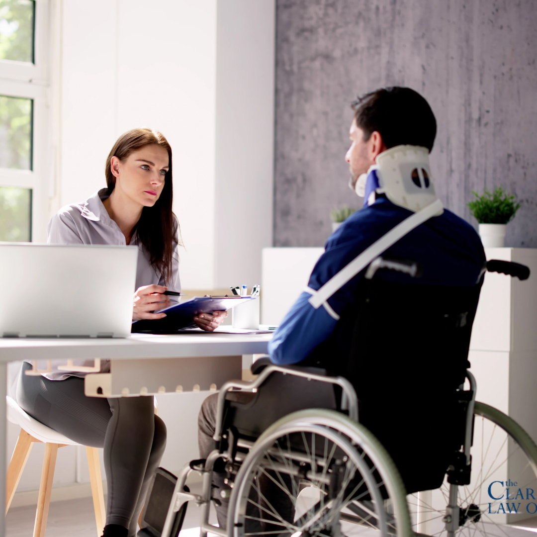 A woman with a clipboard sits at a desk speaking with a man in a wheelchair who is wearing a neck brace. A woman with a clipboard sits at a desk speaking with a man in a wheelchair who is wearing a neck brace.