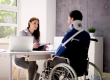 A woman with a clipboard sits at a desk speaking with a man in a wheelchair who is wearing a neck brace.