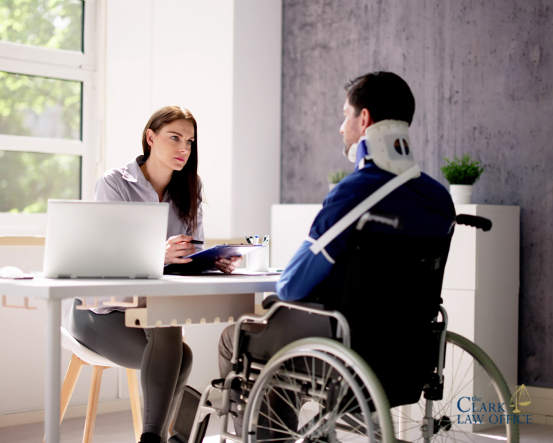 A woman with a clipboard sits at a desk speaking with a man in a wheelchair who is wearing a neck brace. A woman with a clipboard sits at a desk speaking with a man in a wheelchair who is wearing a neck brace.