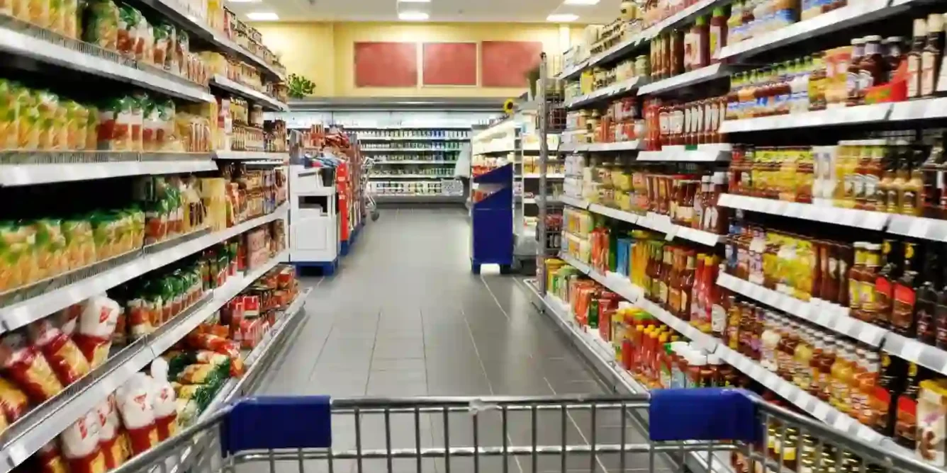 A shopping cart is pushed down an aisle in a well-lit supermarket, with shelves stocked with various packaged food items on both sides. A shopping cart is pushed down an aisle in a well-lit supermarket, with shelves stocked with various packaged food items on both sides.