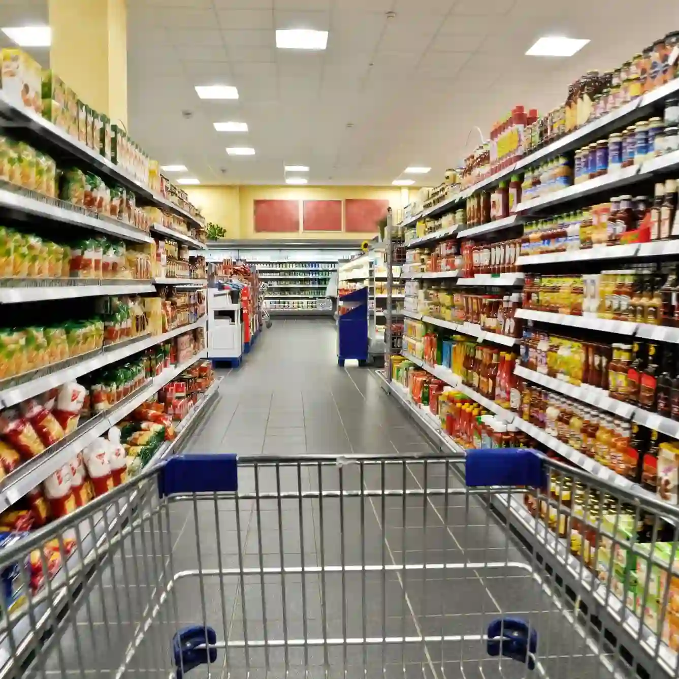 A shopping cart is pushed down an aisle in a well-lit supermarket, with shelves stocked with various packaged food items on both sides.