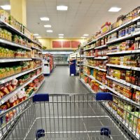 A shopping cart in a grocery store aisle, surrounded by shelves stocked with various food products and condiments.