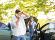 A man stands by two cars with damage, talking on his phone and looking stressed, while a woman stands near one open car door with a raised hood.